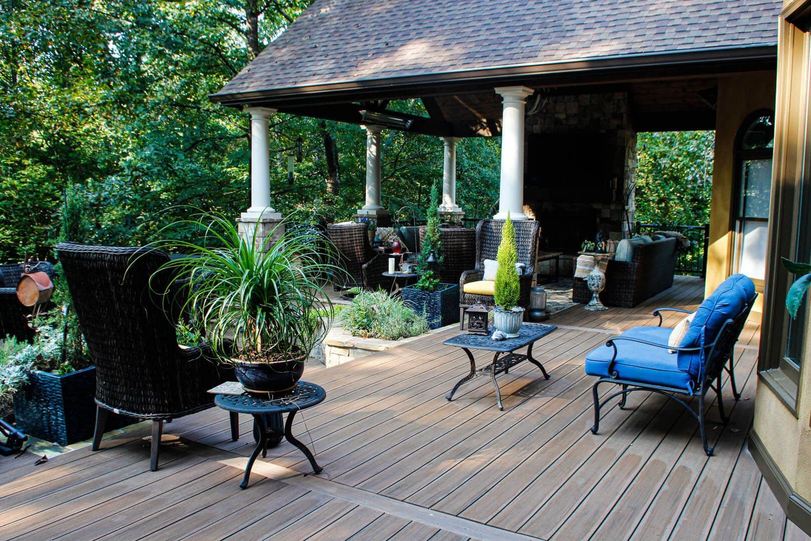 A brown deck with dark patio furniture and a covered area in the background in a wooded yard.