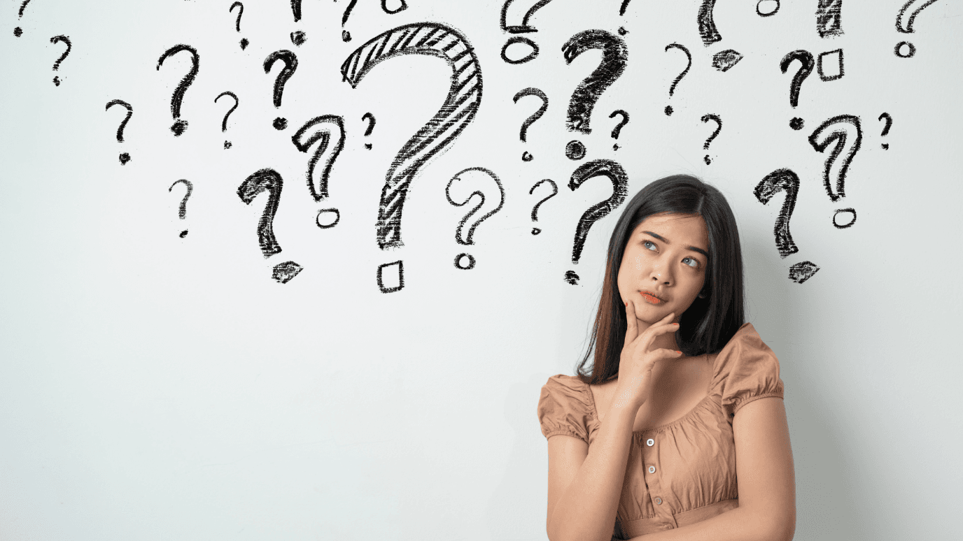 A young woman in a brown top ponders, surrounded by hand-drawn question marks on a whiteboard. She appears thoughtful and curious.