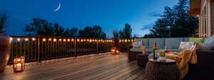 A brown backyard deck at dusk with lighting, patio furniture, and the moon showing.